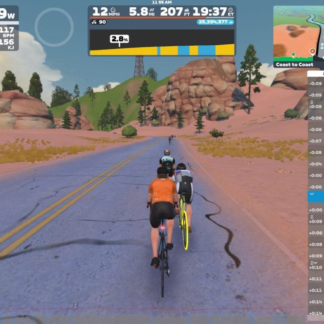 A digital cycling scene shows a group of cyclists riding on a road surrounded by rocky and hilly terrain. The cyclists are in a virtual environment, with stats displayed on the screen, including speed, distance, and power output. The landscape features pink earth and scattered trees.