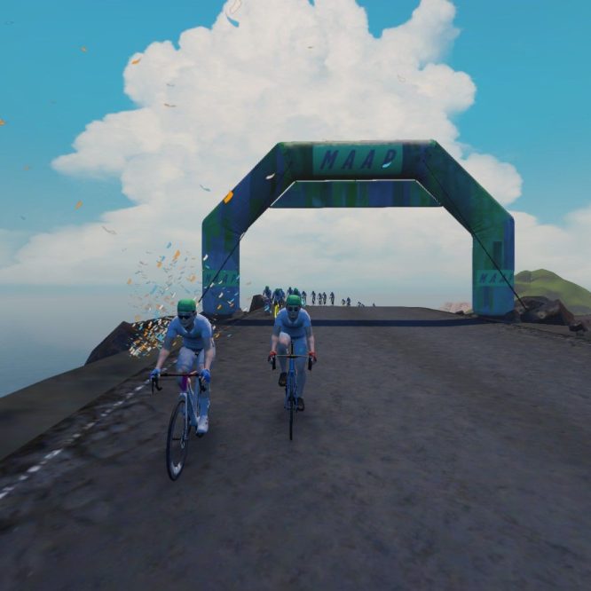 A group of cyclists dressed in light blue uniforms rides toward a large green and blue archway labeled "MAAD." The scene depicts a coastal road with water on one side and a clear blue sky with clouds above. Some confetti is scattered around, adding a festive atmosphere.