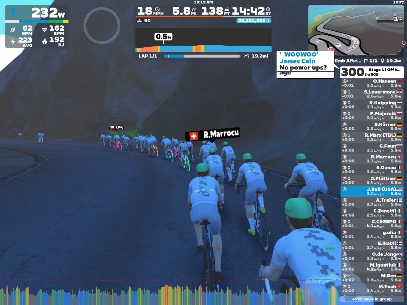 A group of cyclists wearing matching white and green jerseys is riding on a winding road through a canyon-like area. The scene shows several cyclists in a line, with one cyclist in the foreground identifiable by a name tag. The background features rocky terrain, and various performance metrics are displayed on the screen, including speed, distance, and power output.