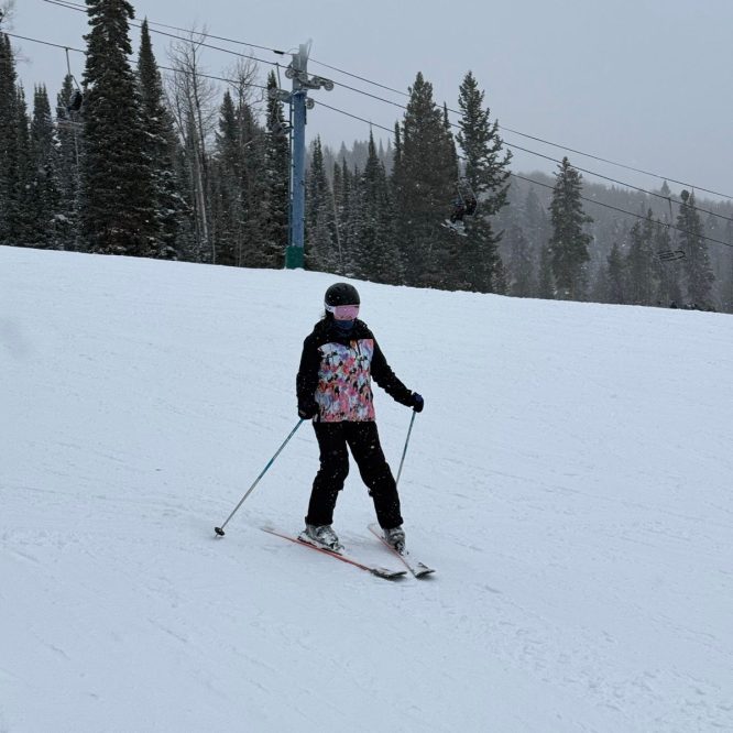 A person skiing on a snowy slope in Logan, UT. The skier is wearing a colorful jacket and black pants, with a helmet and mask. Snow-covered trees and a ski lift are visible in the background.