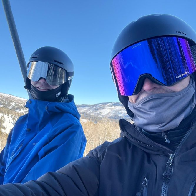 Two people are sitting on a ski lift, smiling and wearing ski helmets and goggles. One person has a blue jacket, while the other is dressed in black. In the background, there are snow-covered mountains and trees under a clear blue sky.