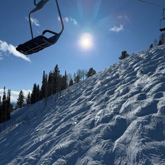 A ski lift chair is suspended above a snowy slope under a bright sun. The hillside features undulating snow patterns and scattered trees in the background, captured against a clear blue sky.