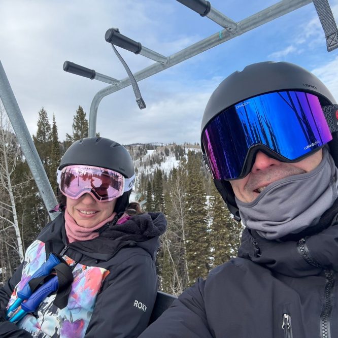 A woman and a man smiling while riding a ski lift. Both are wearing helmets and ski goggles. The background shows snow-covered mountains and evergreen trees under a cloudy sky.