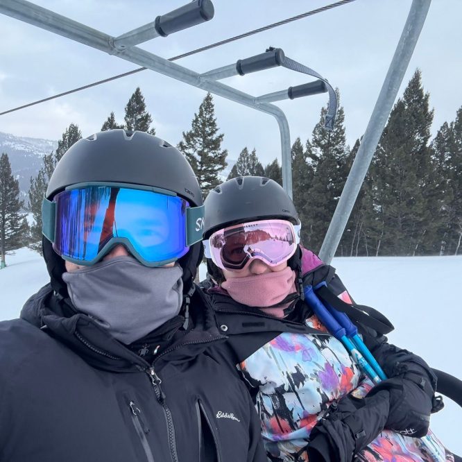 A man and a woman wearing winter skiing gear, including helmets and goggles, sit on a ski lift amidst a snowy landscape. Pine trees and mountains are visible in the background, under a cloudy sky. The woman is holding ski poles and both are partially covered with face masks for warmth.