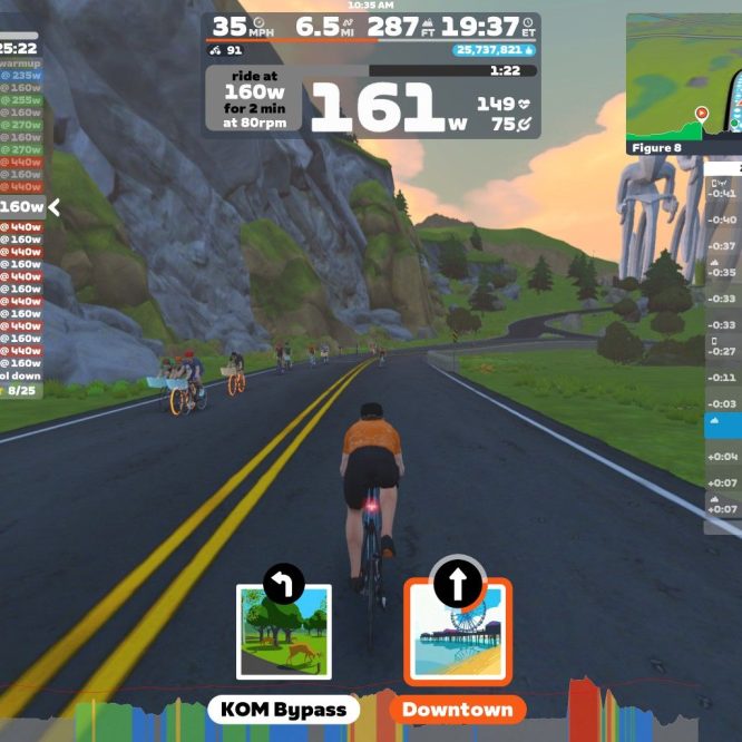 A cyclist in an orange shirt rides on a virtual road in Zwift, a cycling simulation platform. The screen displays performance metrics, including speed, distance, and wattage, along with a leaderboard on the right side showing other riders. Scenic graphics with rocks and trees are visible in the background.