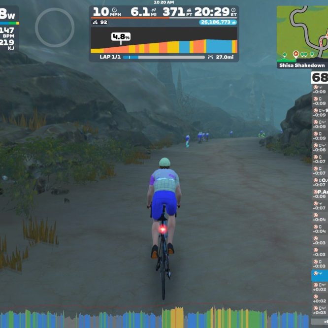 A cyclist navigates a rocky path in a virtual environment on Zwift, with a landscape featuring steep rocks and sparse vegetation. The screen shows performance metrics, including power output, speed, heart rate, and lap information, along with a leaderboard of other participants.