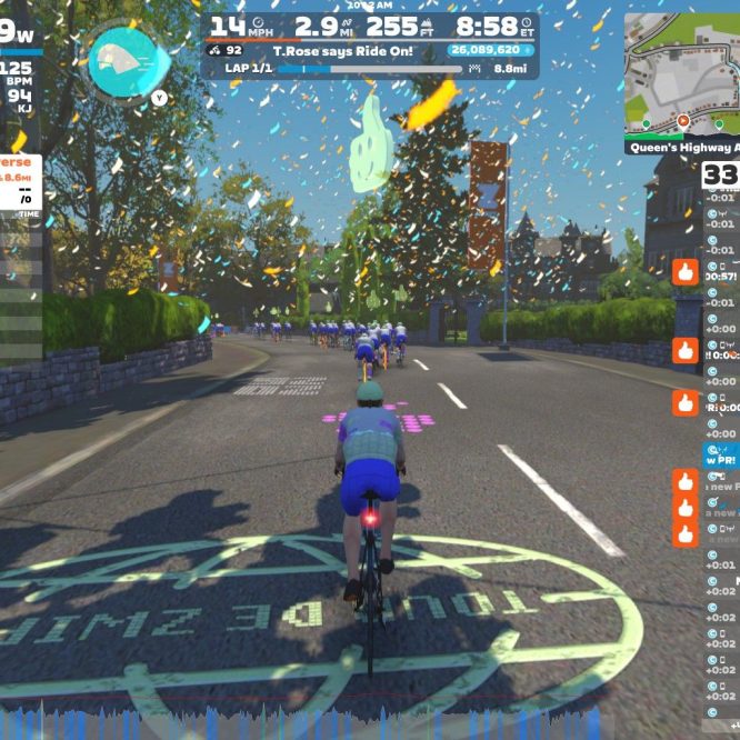 A cyclist is riding on a virtual road in Zwift, surrounded by other riders. The scene is bright and festive, with confetti falling from the sky. The interface shows performance metrics, including power output, heart rate, and speed, along with a leaderboard on the side. A map is visible in the upper corner, and the cyclist is approaching a turn on the course.