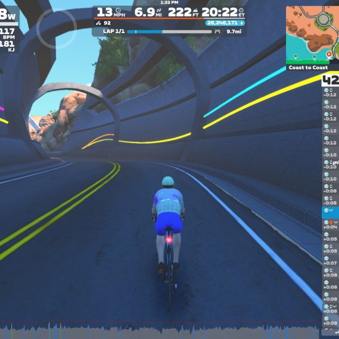 A cyclist is riding through a virtual tunnel in a gaming application, Zwift. The rider is wearing a light blue and white outfit and is surrounded by colorful lines on the tunnel walls. The display shows various performance statistics, including power output, speed, and distance. A map and leaderboard are visible on the right side of the screen. The scene captures a dynamic and immersive cycling experience in a digital environment.