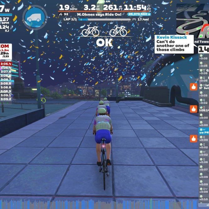 A cyclist rides on a virtual roadway in Zwift, surrounded by floating confetti and digital stats displayed on the screen. The cyclist is part of a leaderboard showing rankings and times for a climbing event called "ROOFTOP KOM." The image includes various performance metrics like power, speed, and heart rate, along with comments from other participants.