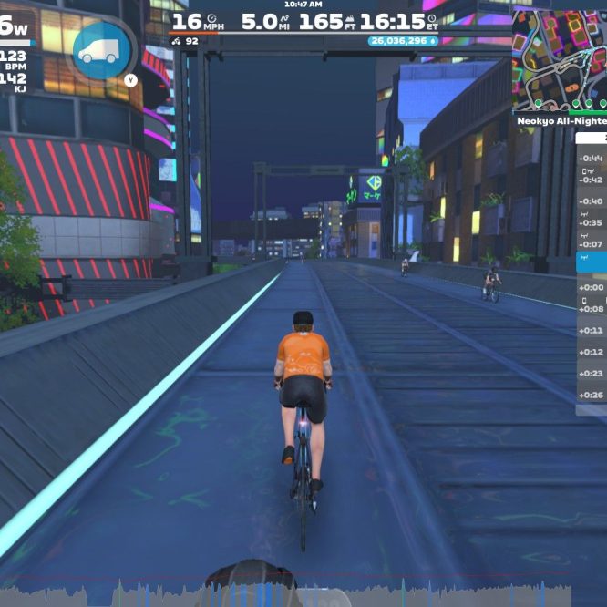 A cyclist in an orange shirt rides on a virtual road in Zwift, surrounded by neon-lit buildings at night. The screen displays various performance metrics, including power output, speed, and heart rate, along with a list of other cyclists nearby. A map of the virtual location is shown in the upper right corner.