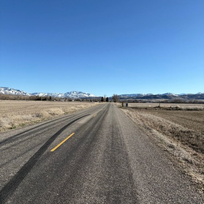A straight, empty road stretches into the distance under a clear blue sky, surrounded by fields and mountains in the background. The landscape is mostly dry, with some patches of snow visible on the mountains.