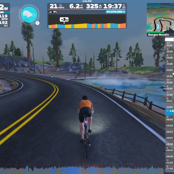 A cyclist in an orange jersey rides along a winding road in a virtual landscape with trees and a body of water. The screen displays various performance metrics, including speed, power, and heart rate, as well as a mini-map and nearby cyclists. The environment has a cloudy sky and rocky terrain.