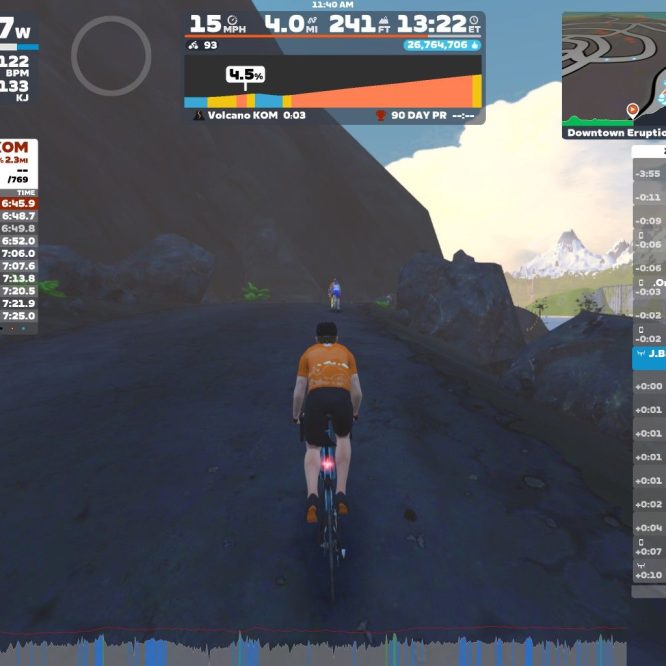 A cyclist in an orange jersey rides on a virtual road within the Zwift platform, which displays performance metrics. The screen shows a leaderboard with live results for the Volcano KOM event, including ranks and times, as well as nearby riders. In the background, a scenic mountainous landscape is visible.