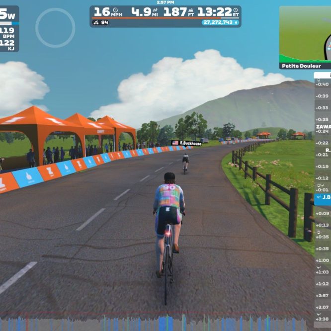 A virtual cycling scene in Zwift, showing two cyclists on a winding road. The path is lined with cheering spectators and orange tents. The sky is blue with some clouds, and a mountainous background can be seen. Performance data and nearby cyclists' information are displayed on the right side of the image.