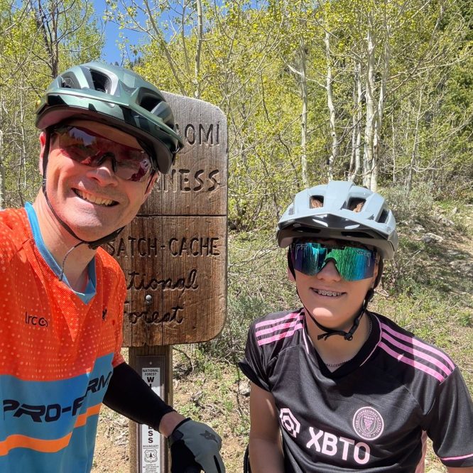 A man and a young girl wearing helmets and sunglasses pose for a selfie at a trail sign in Green Canyon, Utah. The sign indicates the area is part of the Wasatch-Cache National Forest, surrounded by lush greenery.