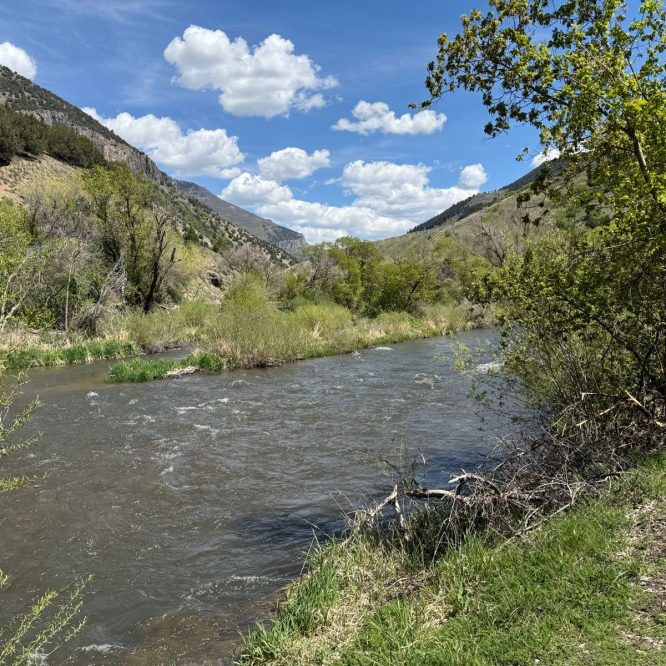 A scenic view of a river winding through lush greenery in Paradise, Utah, with mountains in the background and a partly cloudy sky.