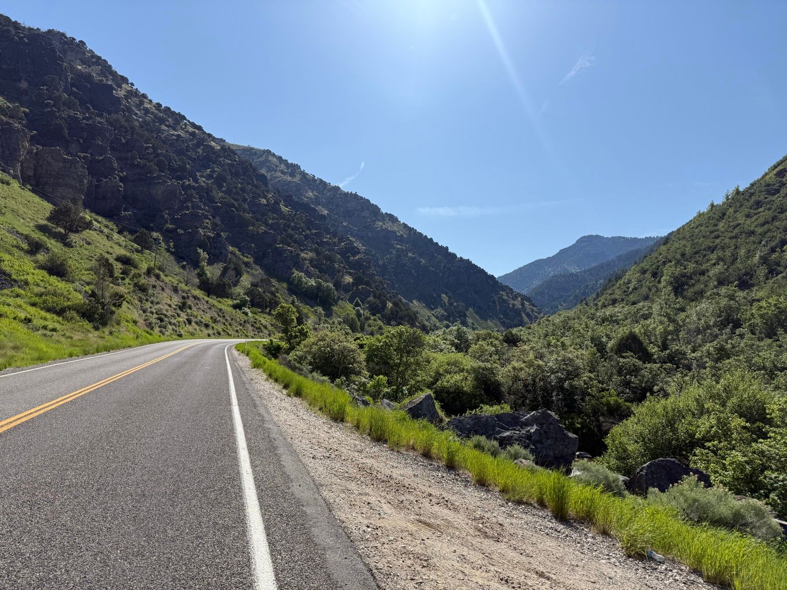 A winding road runs through a green valley surrounded by rocky mountains under a clear blue sky.