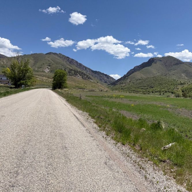 A wide, paved road stretches through a rural landscape in Nibley, Utah. On the left, there is a white house surrounded by trees, and in the background, rolling green hills rise under a bright blue sky dotted with white clouds. Lush green grass lines the road, creating a peaceful, scenic view.