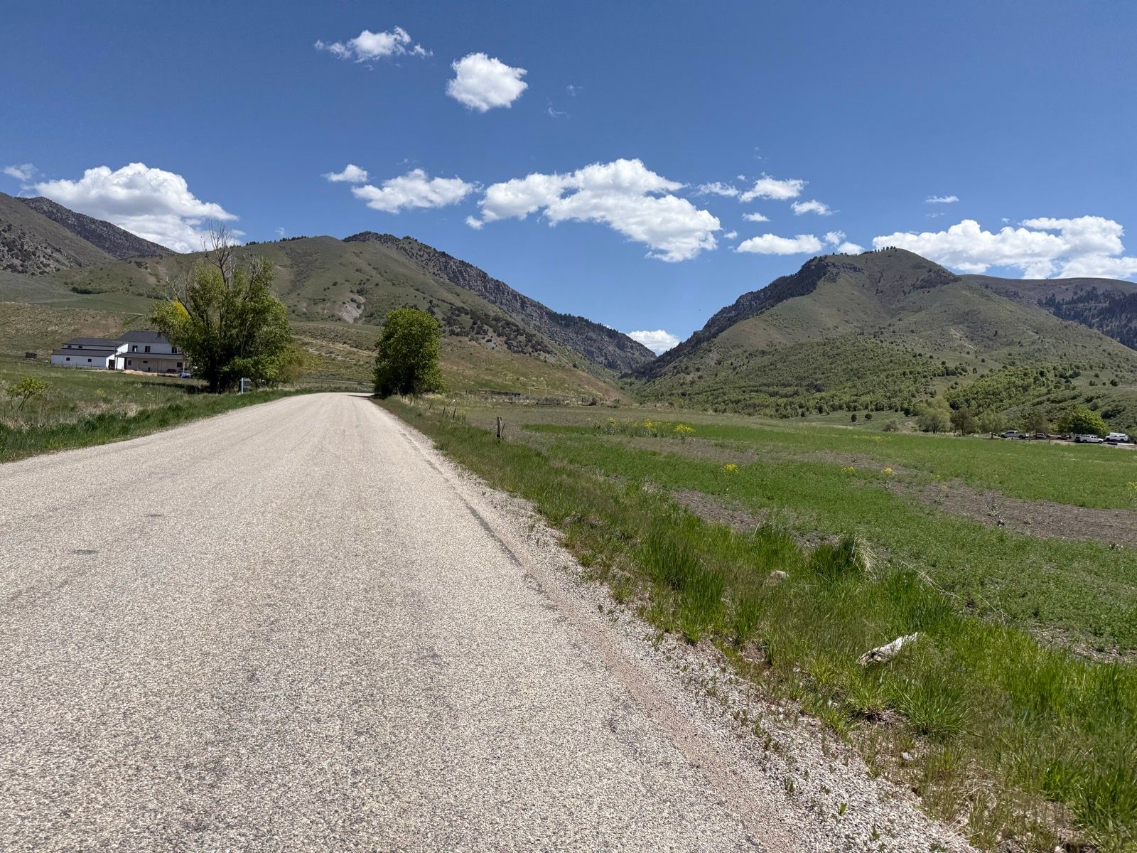A wide, paved road stretches through a rural landscape in Nibley, Utah. On the left, there is a white house surrounded by trees, and in the background, rolling green hills rise under a bright blue sky dotted with white clouds. Lush green grass lines the road, creating a peaceful, scenic view.