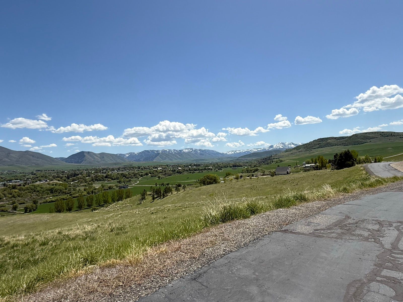 A panoramic view of a green landscape in Nibley, Utah, featuring rolling hills, fields, and distant snow-capped mountains under a clear blue sky with fluffy white clouds. A winding road is visible in the foreground, leading into the scenery.