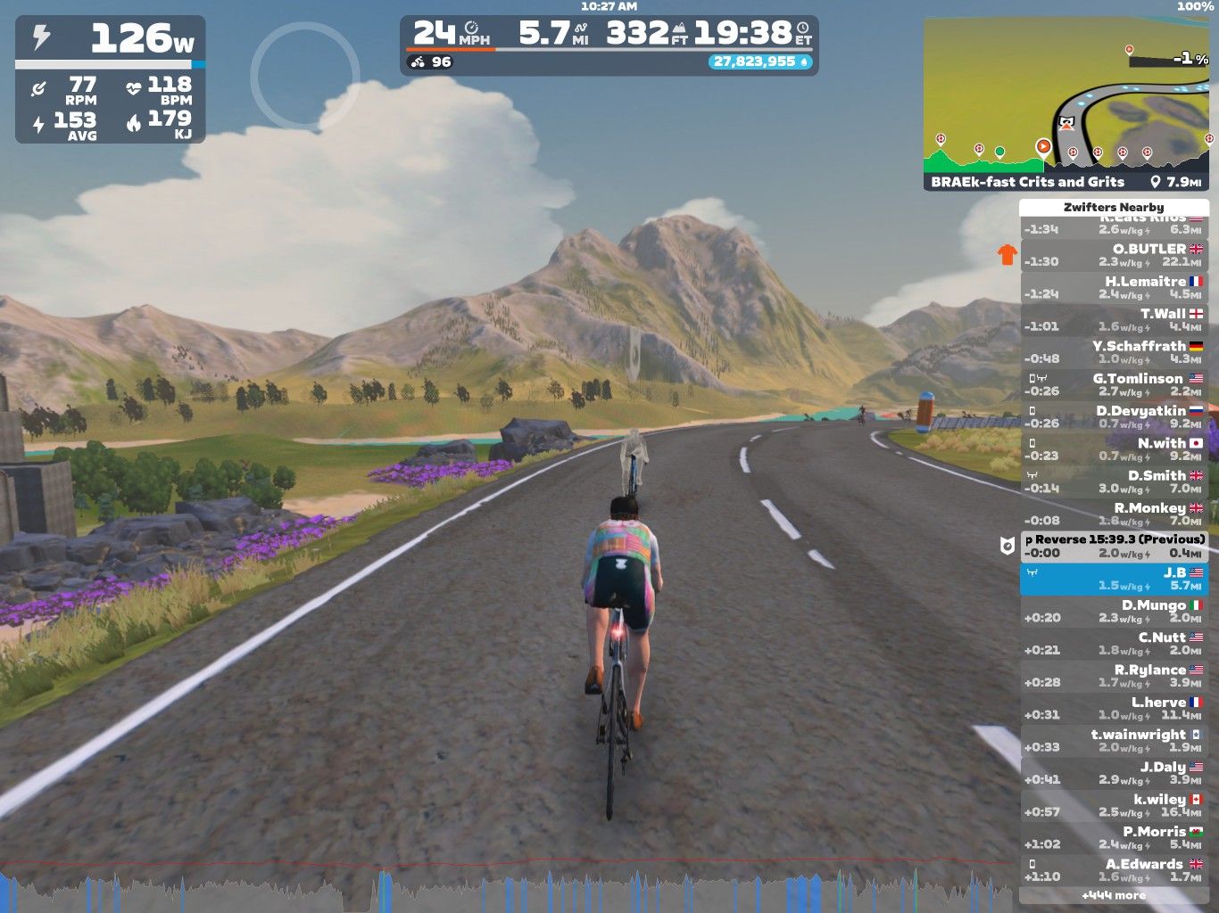 A virtual cycling scene in Zwift, showing a cyclist riding on a paved road surrounded by mountains and greenery. The cyclist is wearing a colorful jersey, and various stats like power output, speed, and heart rate are displayed on the screen. A leaderboard on the side shows other participants' performance metrics.