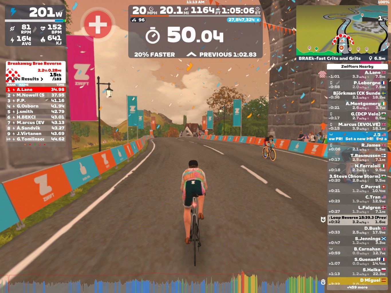 A cyclist is riding on a virtual road in Zwift, displaying live results and statistics on the screen. The scene features colorful banners and a landscape with trees and buildings. The cyclist is in a racing position, and the interface shows details like power output, speed, and rankings.
