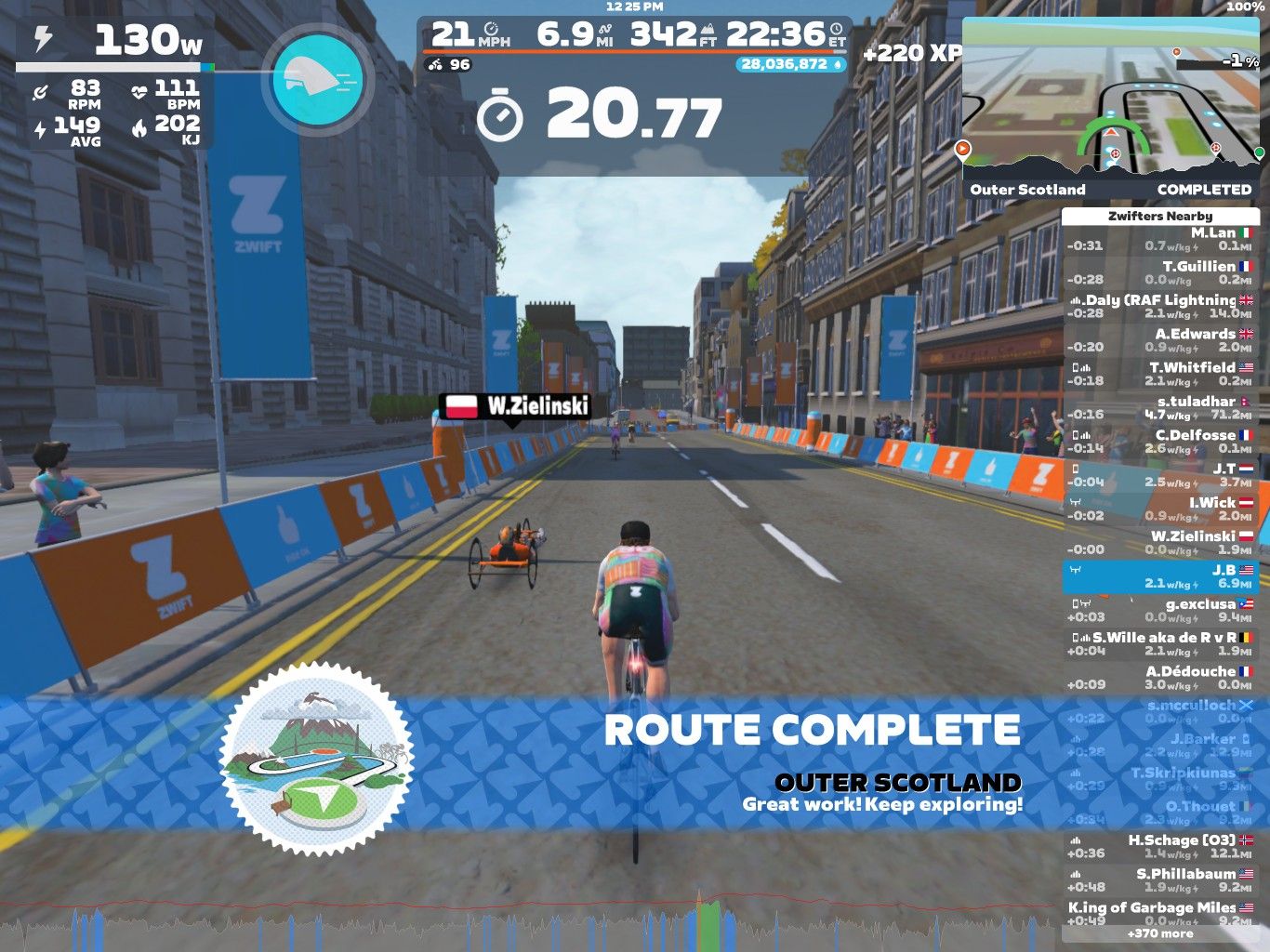 A screenshot from the Zwift cycling game, showing an animated street scene in Outer Scotland. A cyclist with the name