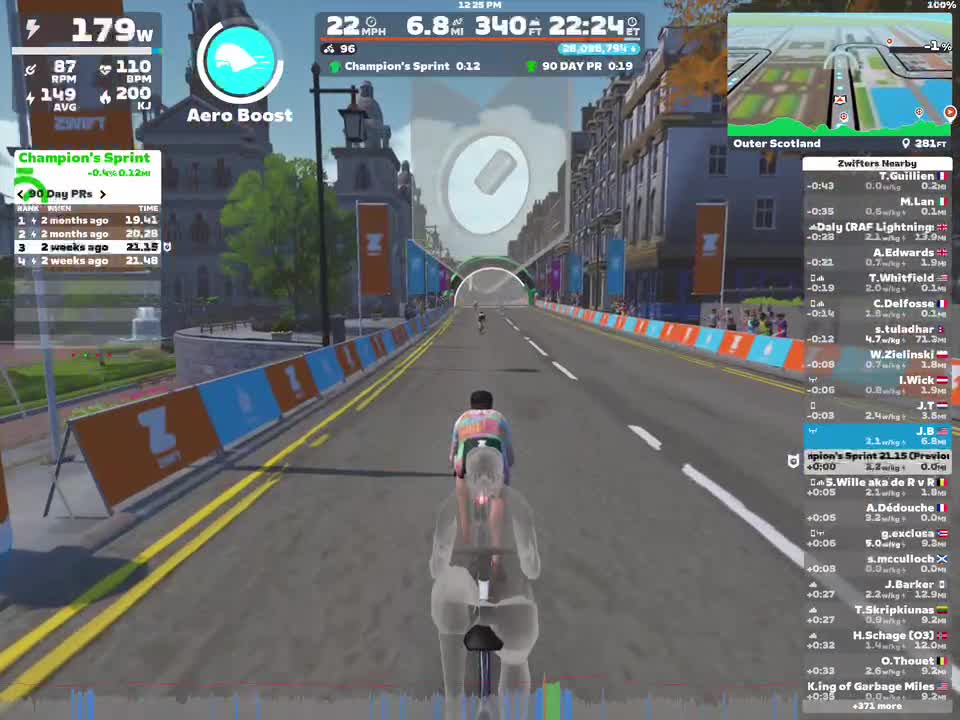 A cyclist is participating in a virtual race in Zwift, shown on a road in a digital depiction of Outer Scotland. The screen displays performance metrics, including power output, speed, and information about nearby cyclists, alongside a leaderboard for a sprint event. The environment features urban buildings and colorful banners along the course.