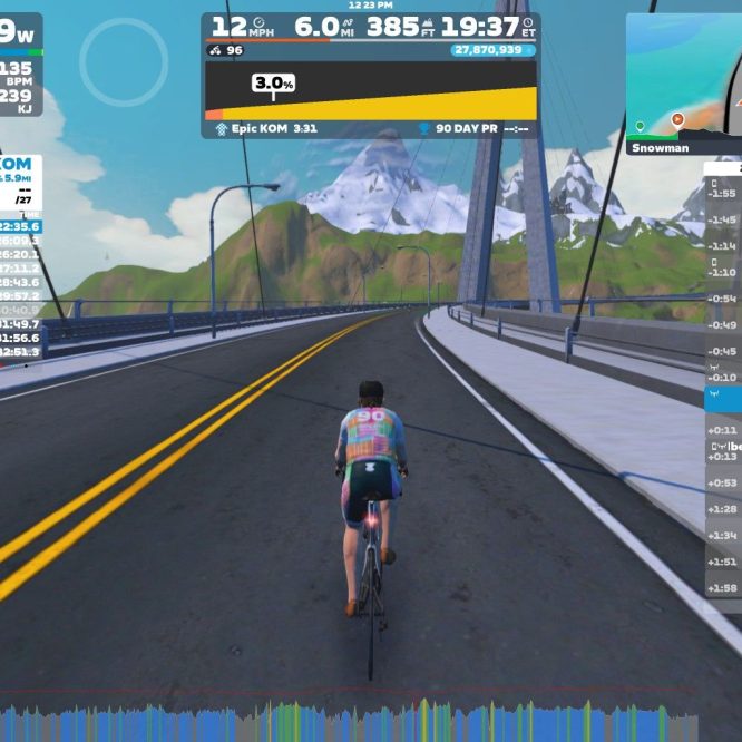 A cyclist is riding on a virtual road in Zwift, surrounded by mountains and a clear blue sky. The screen displays performance metrics, including power output, speed, and elevation, along with a leaderboard showing live results of other riders.