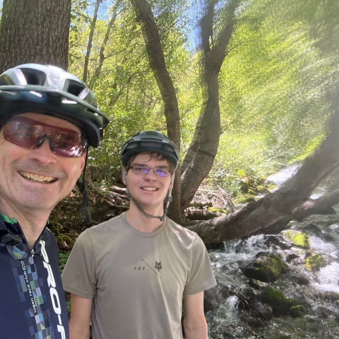 A man and a young adult are smiling for a selfie beside a flowing stream in a lush, green forest. Both are wearing bicycle helmets. Sunlight filters through the trees, creating a bright and cheerful atmosphere.