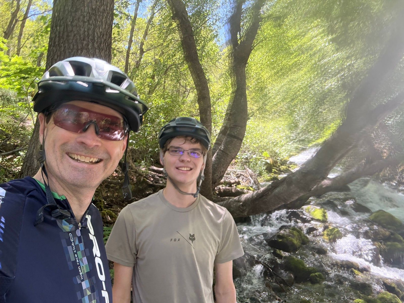 A man and a young adult are smiling for a selfie beside a flowing stream in a lush, green forest. Both are wearing bicycle helmets. Sunlight filters through the trees, creating a bright and cheerful atmosphere.