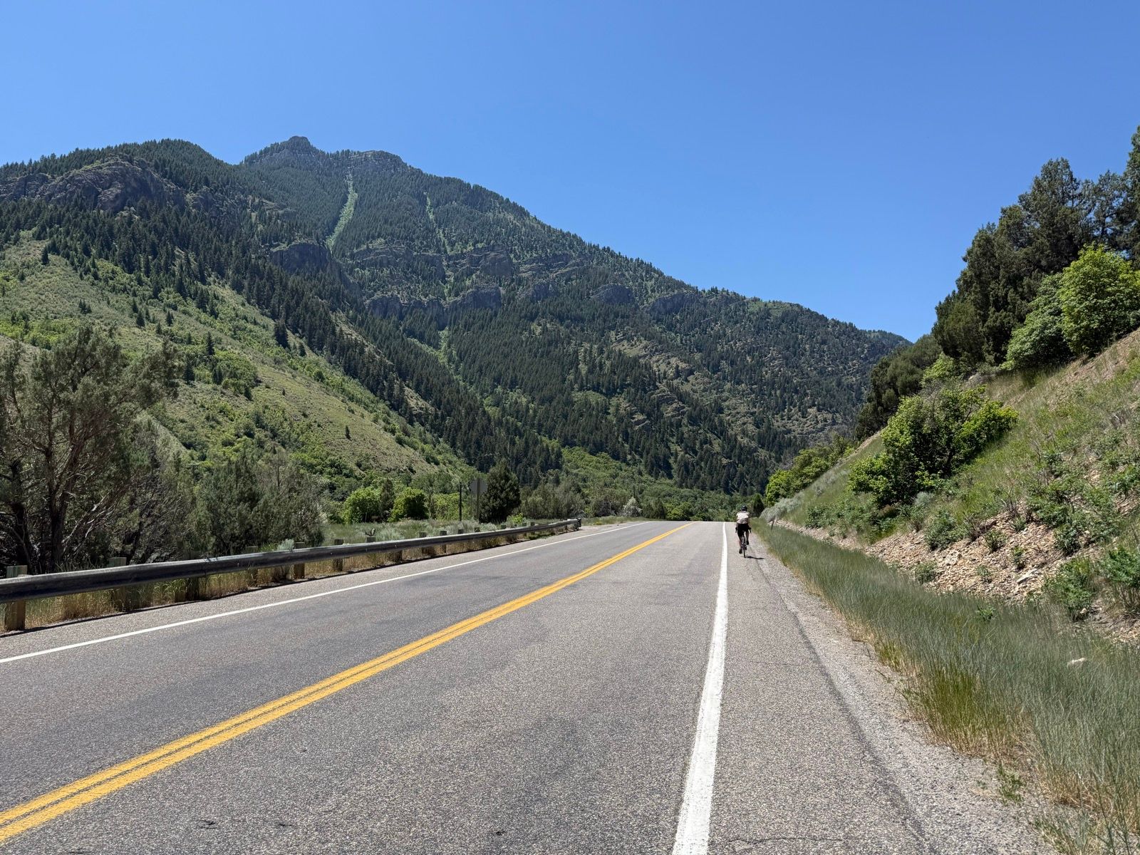 A cyclist rides along a two-lane road in Providence, Utah, with green hills and mountains in the background under a clear blue sky.