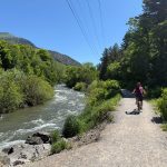 A person rides a bicycle on a gravel path next to a flowing river, surrounded by lush green trees and hills under a clear blue sky in Logan, Utah.