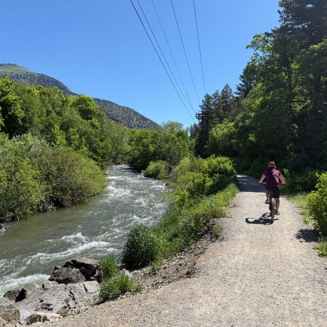 A person rides a bicycle on a gravel path next to a flowing river, surrounded by lush green trees and hills under a clear blue sky in Logan, Utah.