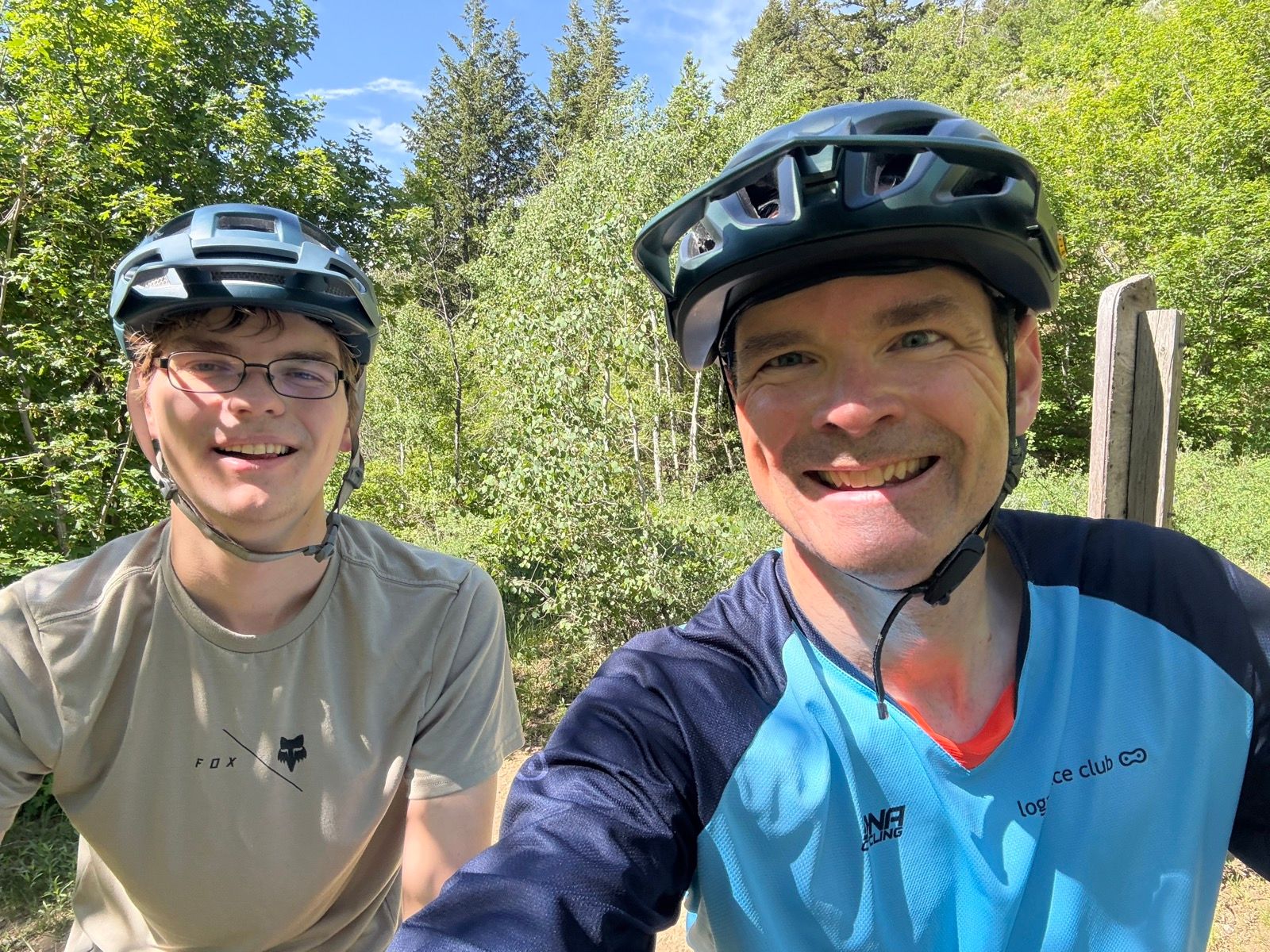 Two people wearing mountain biking helmets pose for a selfie in Green Canyon, Utah. They are smiling in a wooded area with green trees and a blue sky in the background. One is wearing a beige shirt, and the other is in a blue and black jersey.