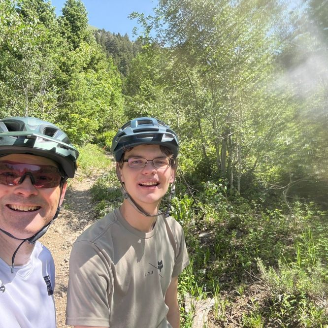 Two people, wearing helmets and sunglasses, are standing on a dirt trail surrounded by greenery in Green Canyon, Utah. The background features trees and sunlight filtering through the leaves.