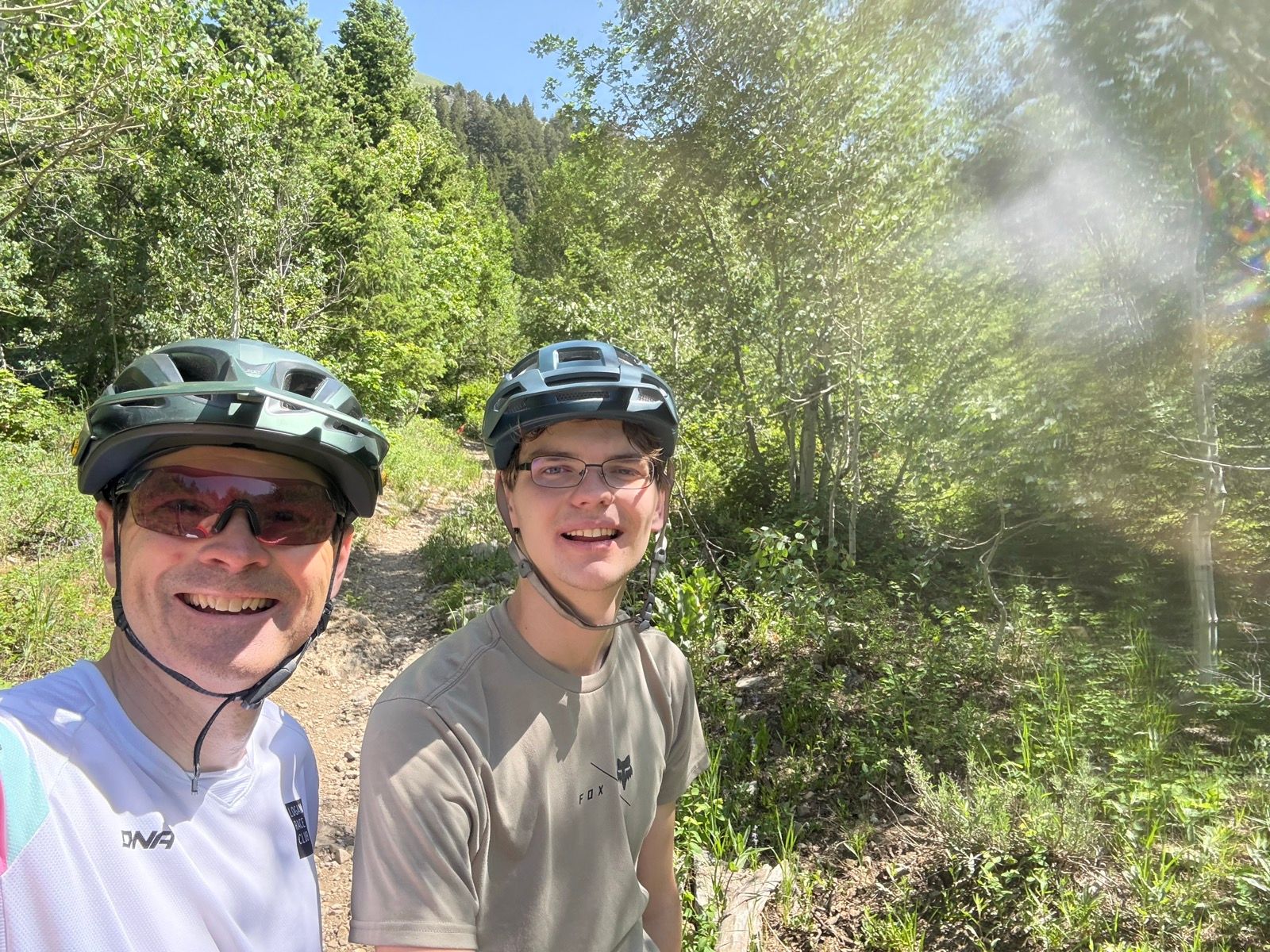 Two people, wearing helmets and sunglasses, are standing on a dirt trail surrounded by greenery in Green Canyon, Utah. The background features trees and sunlight filtering through the leaves.