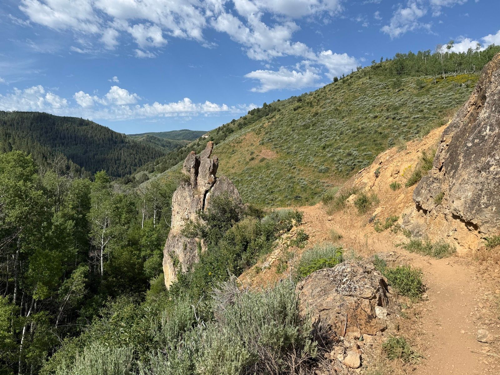 A rocky formation stands prominently on a hiking trail in Wasatch, Utah, surrounded by lush greenery and rolling hills. The blue sky is dotted with white clouds, creating a scenic backdrop.