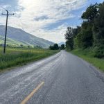 A winding road bordered by green fields and trees, leading towards mountains in the background under a partly cloudy sky at Porcupine Dam, Utah.