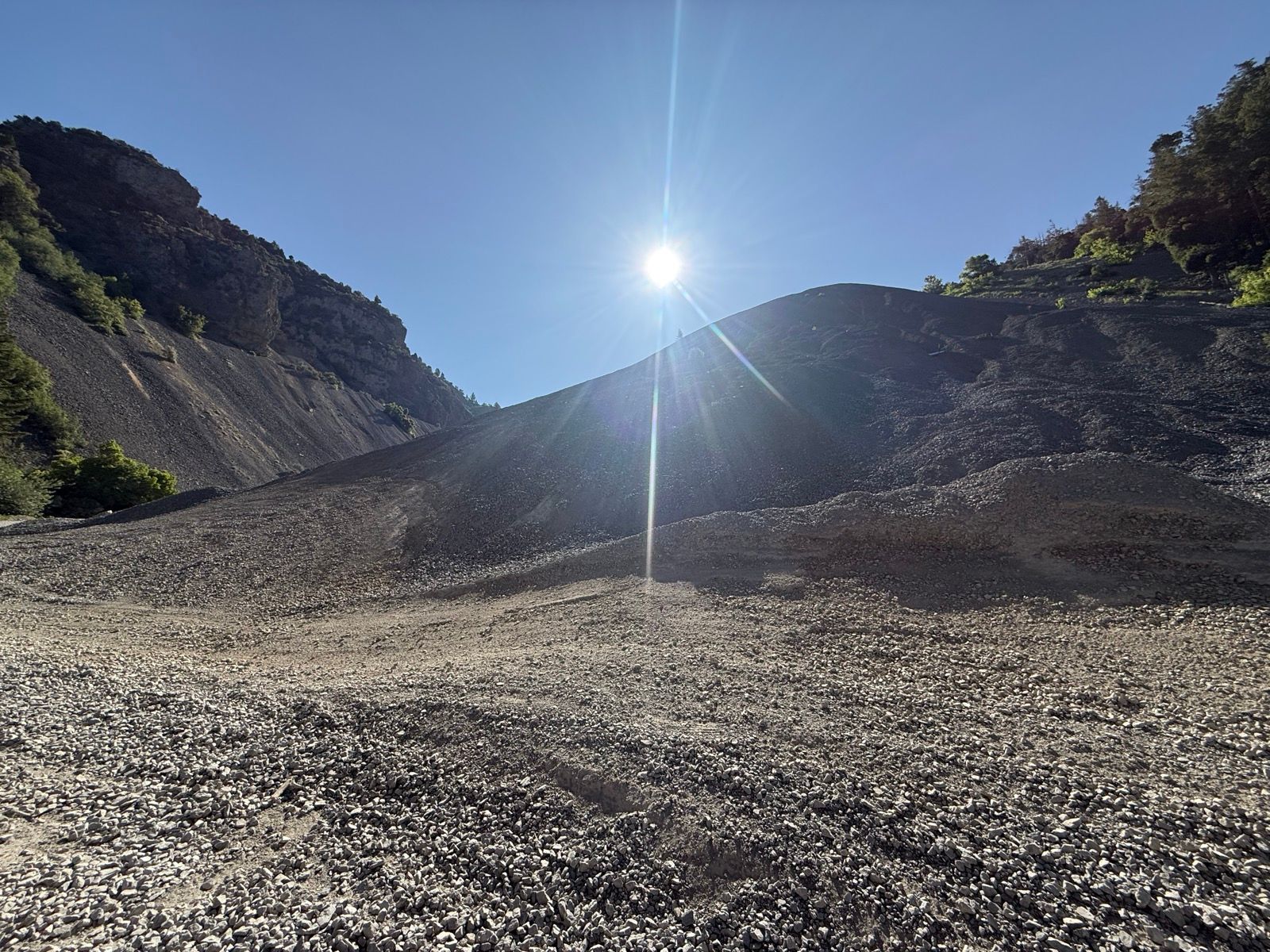 A wide view of Providence Canyon in Utah, featuring a sloping hill of gravel and rocky terrain under a clear blue sky. The sun is shining brightly in the upper right corner. Lush greenery is visible on the hillsides in the background.