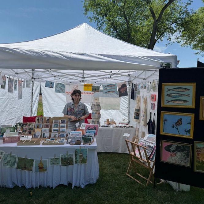 A vendor stands behind a table at an outdoor art fair under a white tent in Providence, Utah. The table is covered with various art prints and greeting cards. In the background, an art display featuring framed pieces hangs on a black panel. The sky is blue with a few clouds, and there are trees in the background.
