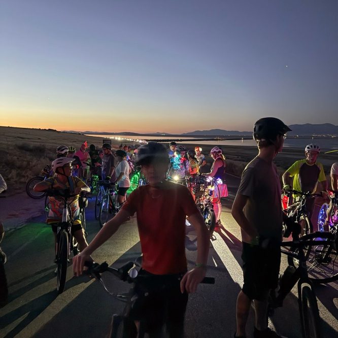 A group of people on bicycles gathers on a road at dusk in Utah. Some riders are wearing helmets, and their bikes are adorned with colorful lights. The sky transitions from orange to deep blue, with distant mountains visible in the background.