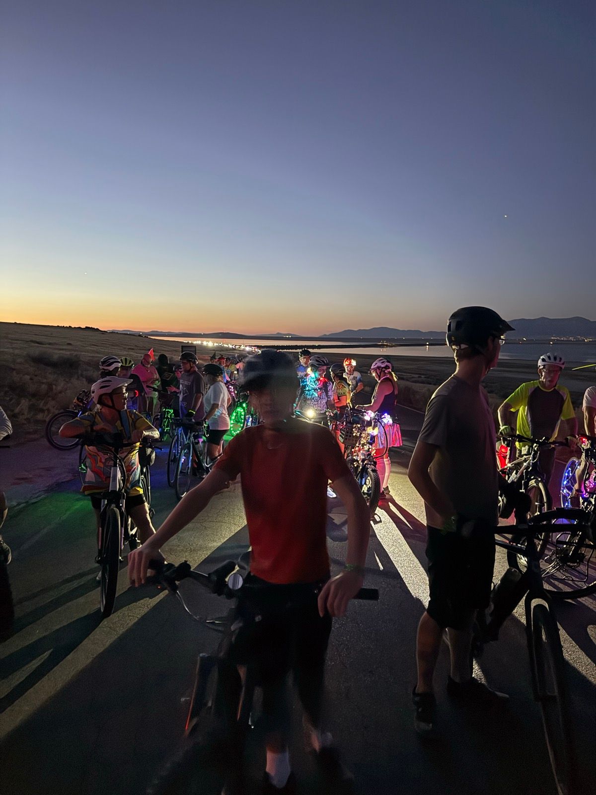 A group of people on bicycles gathers on a road at dusk in Utah. Some riders are wearing helmets, and their bikes are adorned with colorful lights. The sky transitions from orange to deep blue, with distant mountains visible in the background.