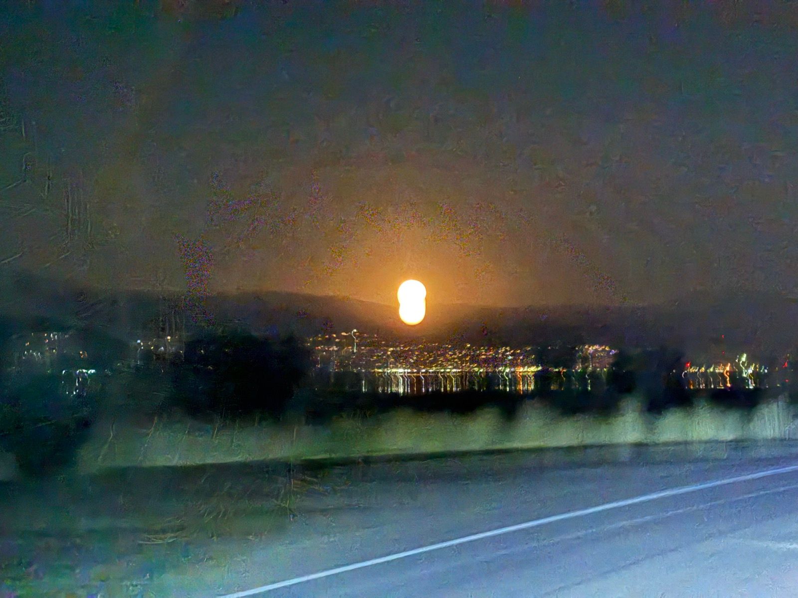 A nighttime view of a bright orange glow on the horizon, likely a sunset or moonrise, illuminating the mountains and reflecting on a body of water. City lights can be seen in the foreground along the roadside.