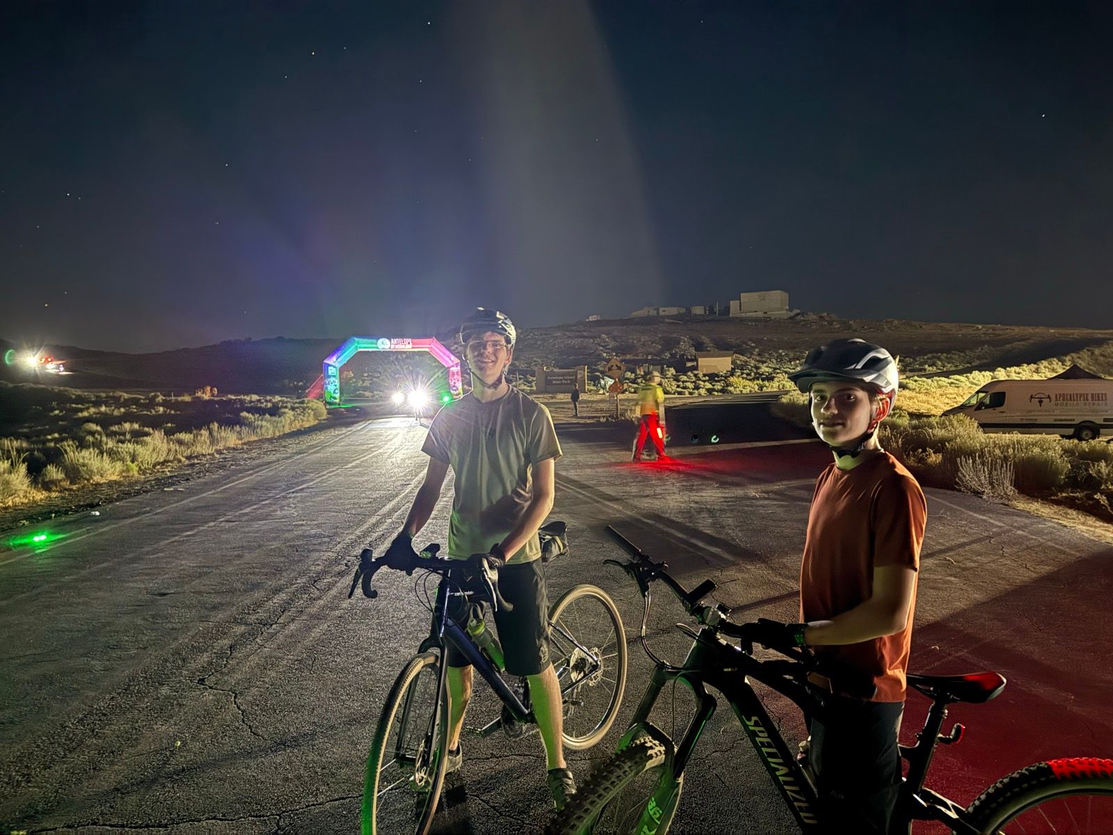 Two young boys stand next to their bicycles on a dark road in Utah, USA. They are wearing helmets and smiling at the camera. In the background, there is a colorful illuminated archway and some distant lights, suggesting an event is taking place. The scene captures a nighttime atmosphere with stars visible in the sky.