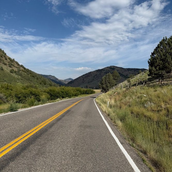 A winding road stretches through green hills under a blue sky with scattered clouds in Nibley, Utah.