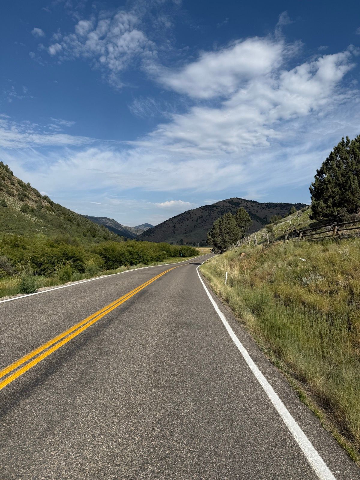 A winding road stretches through green hills under a blue sky with scattered clouds in Nibley, Utah.