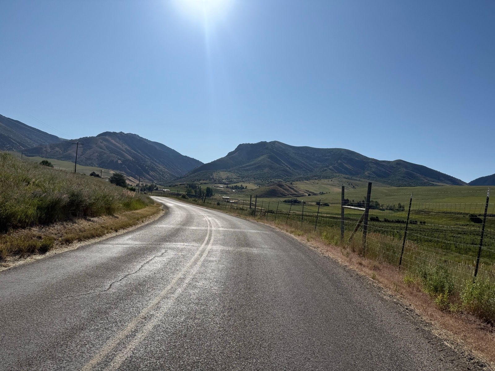 A winding road curves through green fields and hills under a clear blue sky in Nibley, Utah. The landscape features mountains in the background and a fence along the roadside.