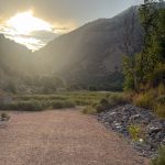 A gravel path in Paradise, Utah, leads towards a view of mountains under a cloudy sky, with the sun setting behind the peaks. Lush greenery and a rocky area are visible on either side of the path.