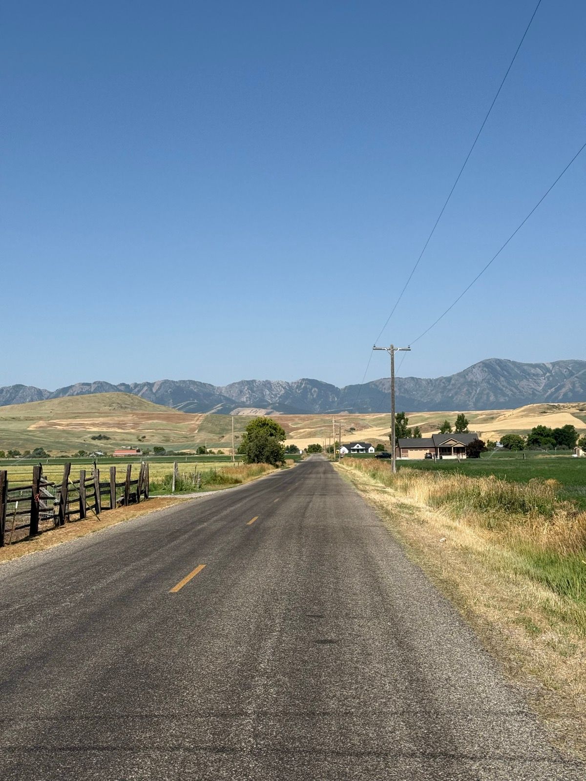 A straight rural road stretches into the distance, flanked by fields and fences. In the background, there are rolling hills and mountains under a clear blue sky. Sparse houses and trees are visible along the roadside.